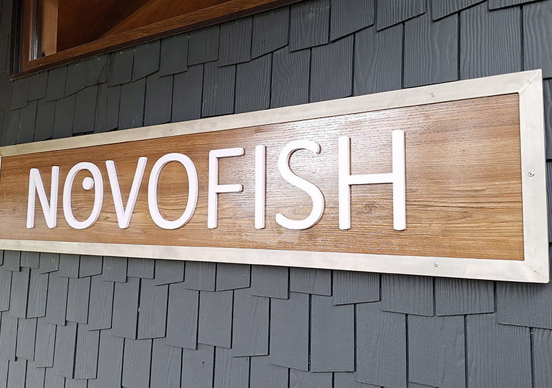 Novofish — Letrero de Alucom revestido con lamina HPL diseño madera, con logotipo volumétrico de PVC espumado y borde de acero inoxidable