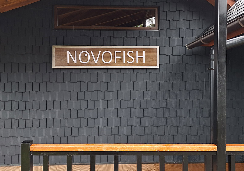 Novofish — Letrero de Alucom revestido con lamina HPL diseño madera, con logotipo volumétrico de PVC espumado y borde de acero inoxidable